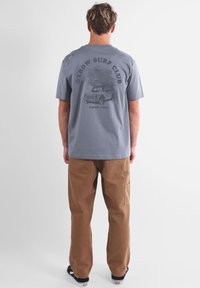 T-shirt en coton gris avec un graphisme sur le thème du surf au dos, associé à un pantalon marron. Le t-shirt a des manches courtes et une coupe décontractée.