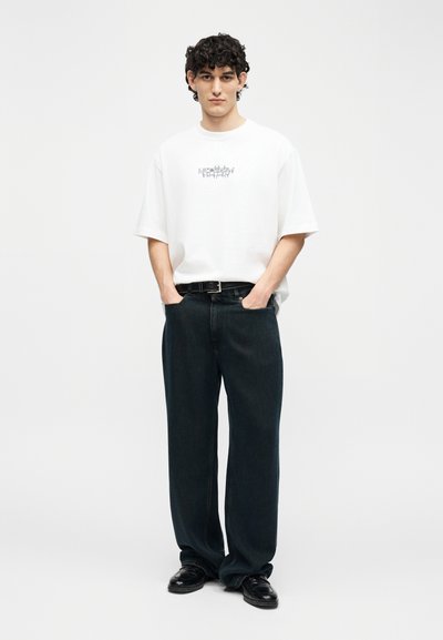 Νεαρός άνδρας με σγουρά μαλλιά, ντυμένος με λευκό φαρδύ t-shirt με μικρό γραφικό, μαύρο τζιν με φαρδιά πόδια και μαύρα παπούτσια, στέκεται με τα χέρια στις τσέπες.