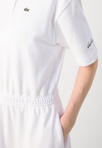 Balta polo suknelė su trumpomis rankovėmis, elastinga juosmens juosta, šoninėmis kišenėmis ir žaliu išsiuvinėtu krokodilo logotipu ant krūtinės. Minkšta audinio tekstūra.