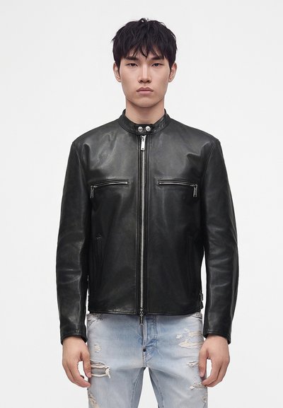 Jeune homme portant une veste en cuir noire zippée avec col pression et un jean bleu clair déchiré, debout devant un fond blanc uni.