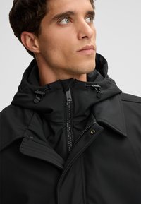 Czarna kurtka z wysokim kołnierzem, podwójnym kapturem, przednim zamkiem błyskawicznym oraz przeszyciami. Gładka tekstura z minimalną ilością elementów metalowych.