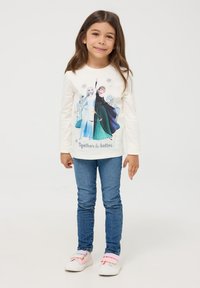 Camicia bianca a maniche lunghe con grafica di due personaggi in abiti colorati, abbinata a jeans blu e sneakers multicolori.