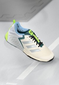 Chaussure de sport avec une tige crème, des accents teal et un logo argenté à trois bandes. Présente une surface texturée et un détail de talon vert pour un soutien.