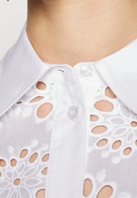 Blouse blanche avec un col cranté ornée de délicats motifs floraux découpés, texture légère et fermeture par un unique bouton.