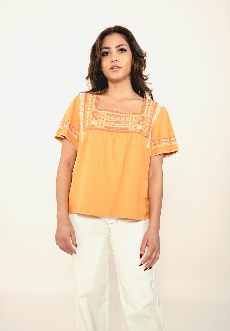 Oranžs kokvilnas tops ar kvadrātveida kakla izgriezumu, īsām piedurknēm un sarežģītu baltu izšuvumu. Apvienots ar baltām platām biksēm.