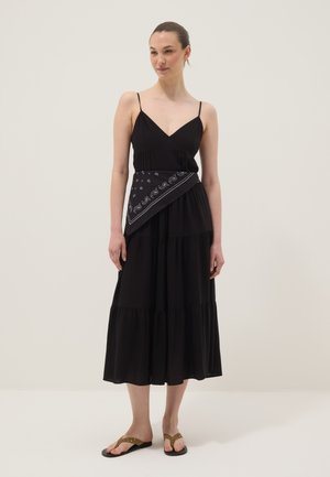 Frau in einem schwarzen ärmellosen Midikleid mit einem Paisley-Muster-Schal, der an der Taille gebunden ist, und braunen flachen Sandalen, steht vor einer schlichten Wand.