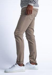 Chinos beige con ajuste tapered, que cuentan con un diseño de cinco bolsillos y puños enrollados, combinados con zapatillas blancas y detalles en verde.