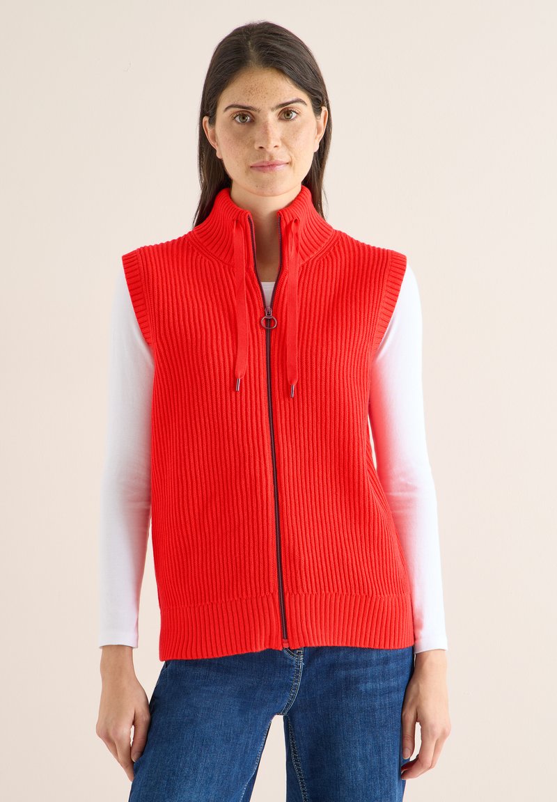 Rotes, geripptes Unterhemd mit hohem Kragen, Frontreißverschluss und Bändern. Getragen über einem weißen Langarmshirt, kombiniert mit blauen Jeans. Glatte Textur.