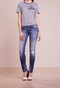 Gestreiftes T-Shirt in Marineblau und Weiß mit dem Text "MEIN UNIVERSE"; ausgewaschene, blaue Skinny-Jeans; wei�ße, hohe Sandalen; minimalistische Gestaltung.