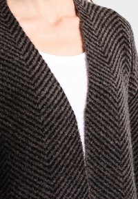 Pletený cardigan v tmavě hnědé barvě s herringbone vzorem, s otevřeným předním dílem a měkkým, texturovaným materiálem.