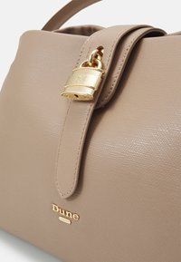 Beige käsilaukku, jossa on teksturoitu keinomateriaali, kultainen lukko ja kultainen brändilogo. Turvallinen läppäsuljin ja sileä hihna.