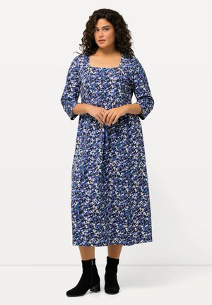 Vestido largo de estampado azul con mangas tres cuartos y escote cuadrado, que presenta formas abstractas en blanco, negro y turquesa. Botines negros hasta el tobillo.