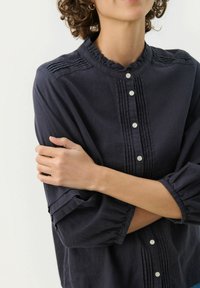 Námořnická modrá lněná košile na knoflíky s volánovým límcem, vertikálními sklady a nařasenými rukávy. Obsahuje bílé knoflíky a jemnou texturu.