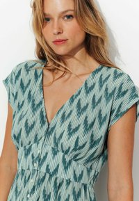 Robe vert menthe avec un décolleté en V profond, manches courtes et un tissu à motifs comprenant des lignes verticales et des textures subtiles, détail de boutons à l'avant.