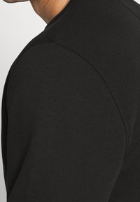 Maglia nera aderente a maniche lunghe con trama a coste, scollo rotondo e dettagli di cuciture visibili sulle spalle e sui lati.