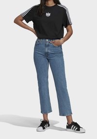 Vrouw in een zwart Adidas T-shirt met witte strepen, blauwe hoge taille jeans en zwart-witte Adidas sneakers, staande met handen op de heupen.