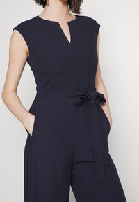 Jumpsuit bez rukávů v námořnické modři s bočními kapsami, výstřihem do V na přední straně a uvázaným látkovým páskem v pase.