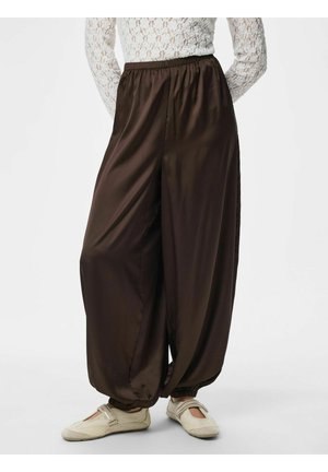 Kvinde iført løse mørkebrune satinbukser med elastisk talje og manchetter, kombineret med beige flade sko og en hvid blonde-top.
