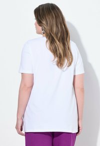 Hvid kortærmet t-shirt med løs pasform, enkelt design og rund hals, båret med lilla bukser, der fremhæver en glat bomuldsstruktur.