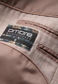 Étiquette de vêtement « ombre QUINTESSENCE M » cousue sur un tissu beige et marron texturé à l’intérieur d’un vêtement.