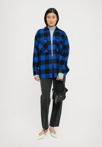 Camicia di flanella a scacchi blu e nera, dolcevita grigio, jeans neri, scarpe bianche a punta e borsa nera strutturata.