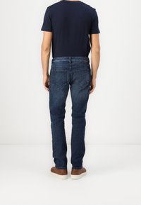 Dark blue denim jeans with a slim fit, featuring two back pockets and a leather patch on the waistband, worn with a navy t-shirt and brown shoes.