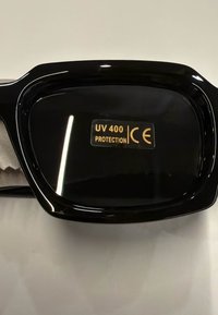 Lente de gafas de sol negras que muestra una etiqueta dorada con "Protección UV 400" y marcado CE sobre un fondo claro.