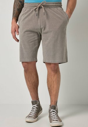 Uomo che indossa pantaloncini grigi con coulisse fino al ginocchio e sneakers alte grigie, in piedi con una mano in tasca e tatuaggio sull'avambraccio sinistro.