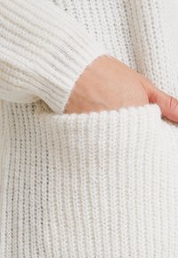 Biały sweter dziergany z fakturą prążkowaną, widoczną kieszenią i miękkim, puszystym wykończeniem. Rękaw ma prążkowany mankiet.