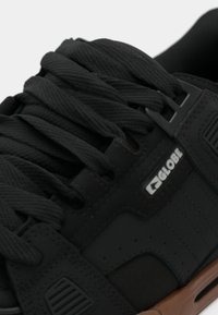 Sorte sneakers i tekstureret materiale, med flade snørebånd, logo-detalje, forstærkede snørehuller og en brun gummi sål.