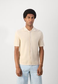 Short-sleeve, cream-colored knitted polo shirt with a textured pattern, button closure, and contrasting collar. Worn with light blue jeans.