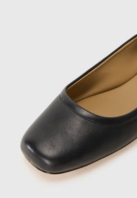Ballerine en cuir noir avec un bout carré arrondi, texture lisse et intérieur rembourré beige, présentant des détails cousus le long de l'ouverture.