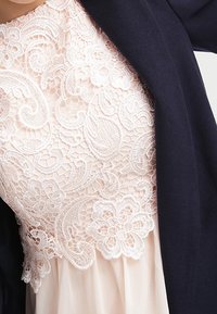 Blazer blu navy abbinato a una maglia rosa chiaro in pizzo floreale con motivi intricati e un tessuto fluido e trasparente sotto.