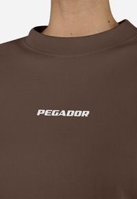 Barna póló magas nyakkal és rövid ujjakkal, fehér "PEGADOR" logóval az elején. Sima szövettextúra.