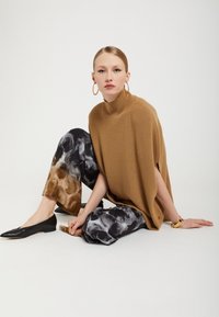 Ruskea neule oversized poncho, jossa on sivuhalkiot, yhdistettynä mustavalkoisiin marmorikuvioisiin leveälahkeisiin housuihin. Mustat loafer-kengät täydentävät asukokonaisuutta.