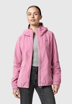 Junge Frau trägt eine hellrosa Kapuzenjacke über einem grauen T-Shirt und schwarzen Jeans, steht vor einem schlichten grauen Hintergrund.