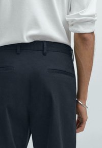 Námořnické kalhoty na míru se zadními kapsami, hladkým materiálem a poutky na opasek. Modelka má na sobě bílé tričko s dlouhým rukávem a stříbrný náramek.