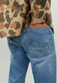 Camisa de camuflaje en tonos marrones y verdes, combinada con jeans de mezclilla azul claro que tienen bolsillos traseros y detalles de costura curva.