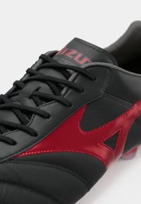 Chuteira de futebol em couro preto com detalhes em vermelho e uma superfície texturizada, apresentando atacadores pretos e um logótipo na língua.