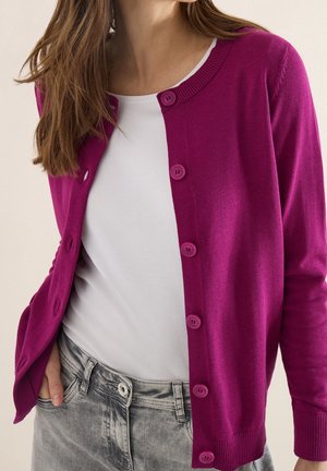 Femme portant un cardigan boutonné magenta vif sur une chemise blanche et un jean gris, une main dans la poche, montrée du cou jusqu'aux hanches.