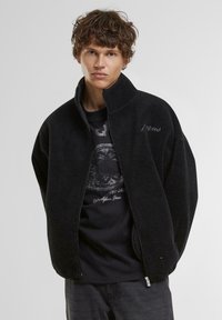 Veste en polaire noire avec col montant, fermeture éclair et poches latérales. Portée par-dessus un t-shirt graphique sombre. Tissu doux et texturé.
