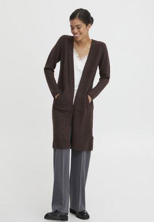 Brauner gestrickter Cardigan mit offenem Vorderteil, langen Ärmeln und aufgesetzten Taschen, kombiniert mit grauen weit geschnittenen Hosen und schwarzen Schuhen.