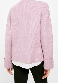 Rückansicht einer Person, die einen lilafarbigen, gestrickten Pullover mit hochgeschlagenen Ärmeln über einem weißen Hemd und schwarzen Hosen trägt.