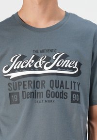 Grå bomull T-skjorte med et stort grafisk design med teksten "Jack & Jones" og "Superior Quality Denim Goods" i kontrasterende farger.
