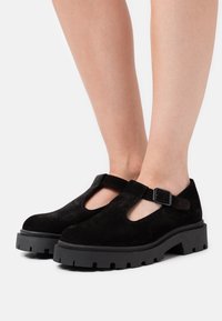 Černé semišové boty s T-páskovým designem, nastavitelnou sponou a chunky podrážkou s gripem pro zvýšenou pevnost a odolnost.