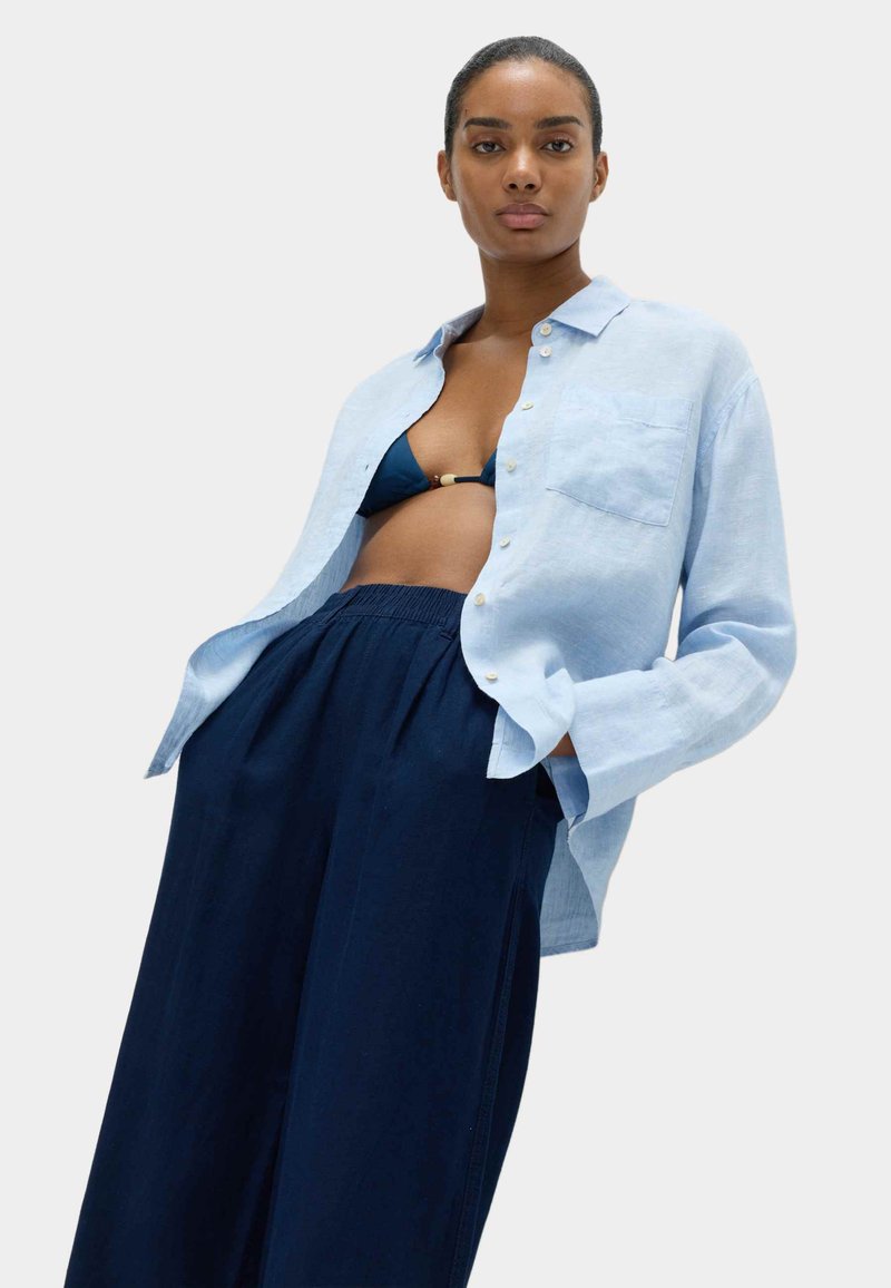 Světlomodrá lněná košile s dlouhým rukávem a předním kapsou, nošená rozepnutá přes tmavě modrý bikini top a tmavě modré široké kalhoty.