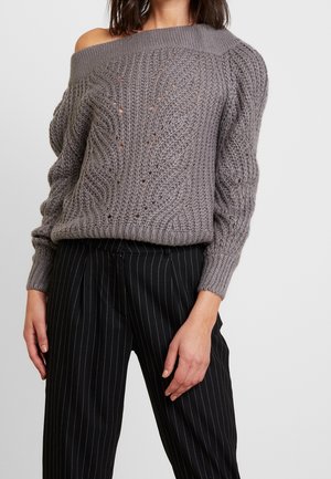 Maglione grigio a spalle scoperte in maglia con motivo a trecce e punti aperti, abbinato a pantaloni neri a righe a vita alta.