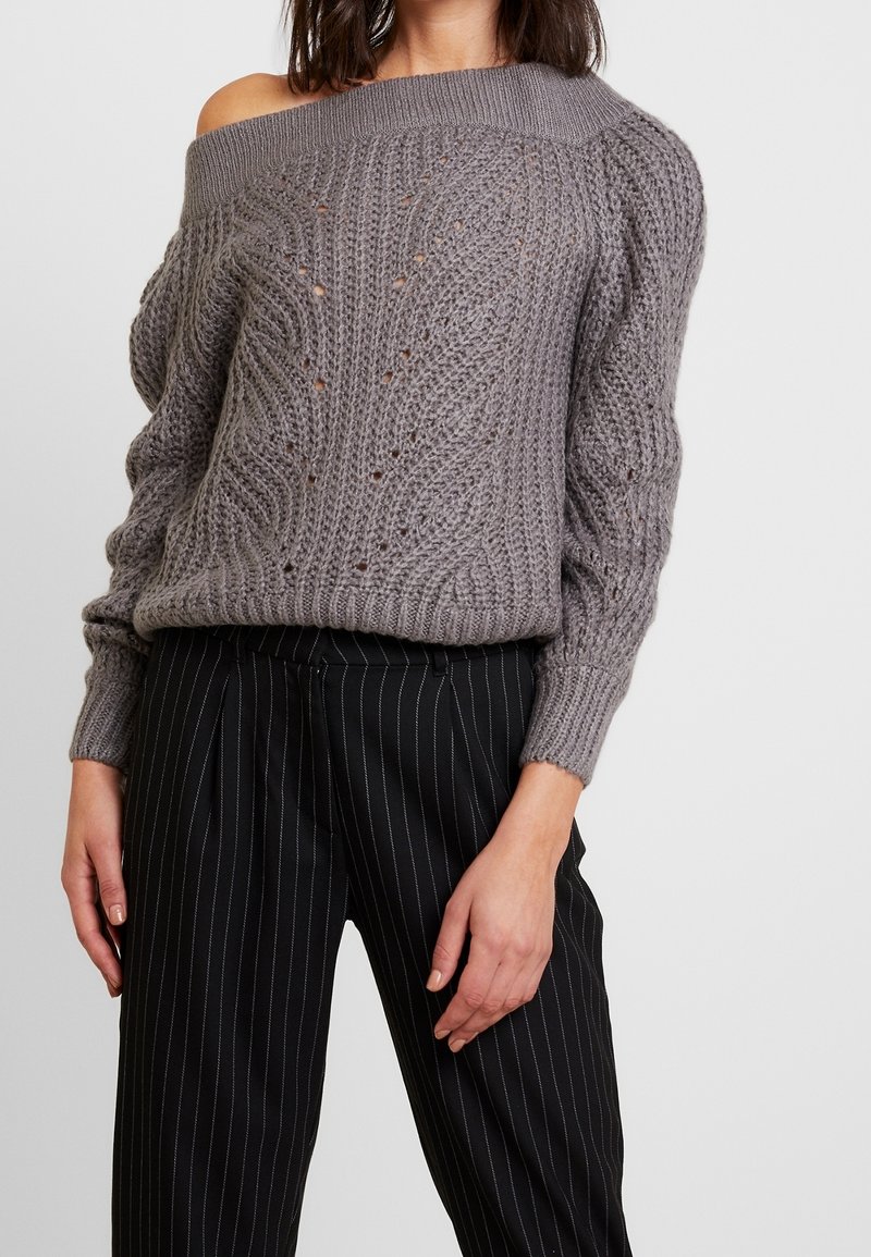 Pull gris en tricot à épaules dénudées avec motif en torsades et mailles ouvertes, associé à un pantalon taille haute à rayures noires.