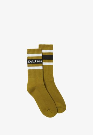 Kaks mustard-keltaista crewsukkia, joissa on valkoisia ja mustia raitoja ylhäällä, toinen sukka esittelee "Dickies"-merkin nimen valkoisella tekstillä.