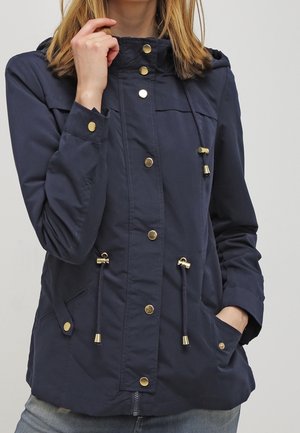 Femme portant une veste à capuche bleu marine avec des boutons dorés et des cordons ajustables, une main dans la poche, l'autre tenant le col.